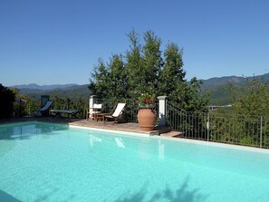 A clear blue swimming pool with lounge chairs and a large planter, surrounded by a metal fence and trees, with mountains in the background.