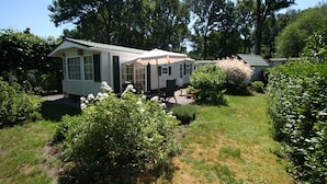 A small, single-story mobile home with a covered porch, surrounded by lush greenery and trees. - Chalet in Stegeren Near Hiking Trails (Stegeren)