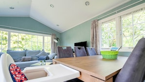 A dining area with a wooden table, chairs, and a high chair. There are windows with curtains, a television, and a basket on the table. - Chalet in Stegeren Near Hiking Trails (Stegeren)