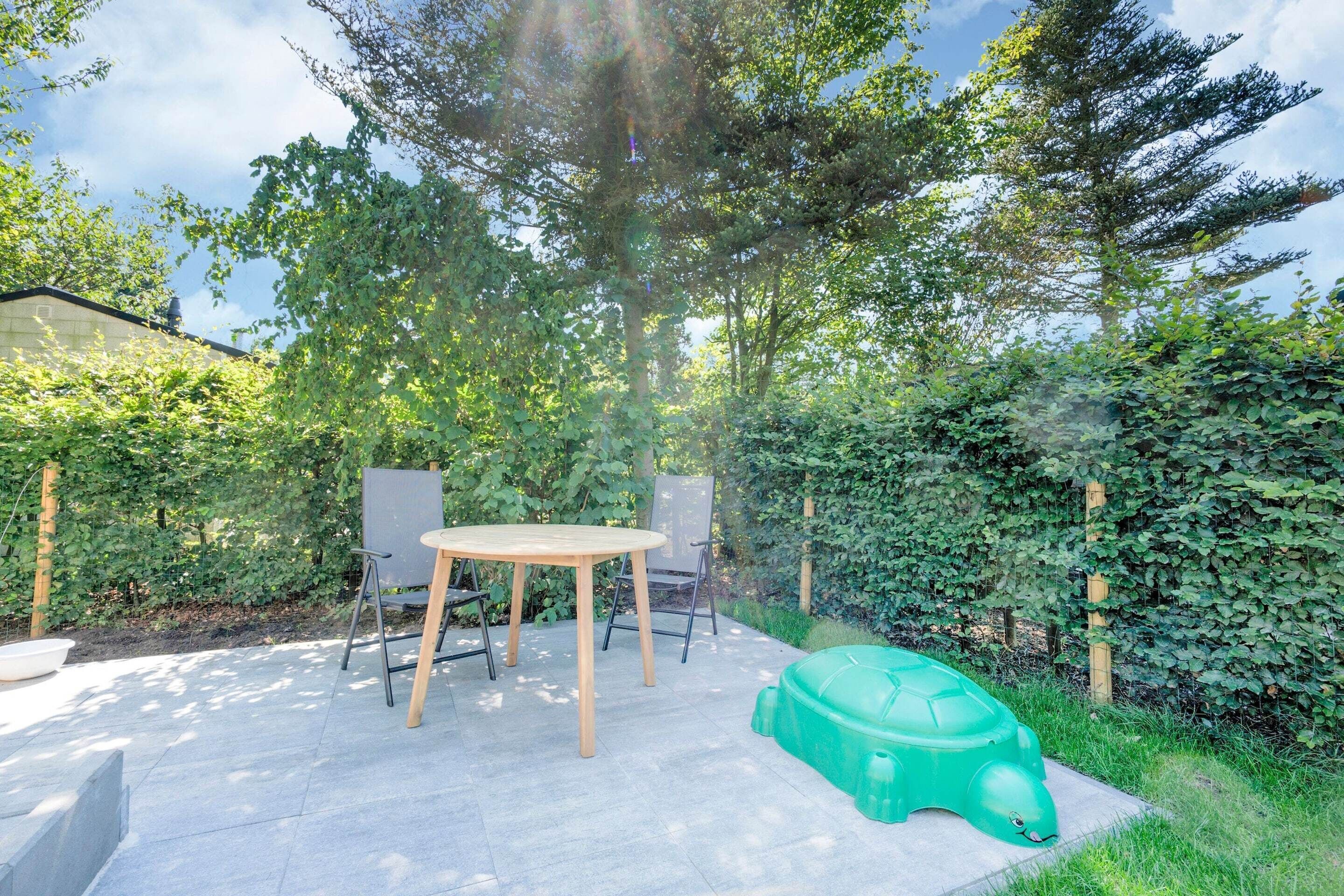 A patio with a round table and chairs, surrounded by lush greenery and a large tree.