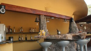Un espace bar avec un mur jaune incurvé, des poutres en bois et des piliers en pierre. On y trouve des tabourets de bar, des étagères remplies de bouteilles et une cloche suspendue à un pilier en pierre.