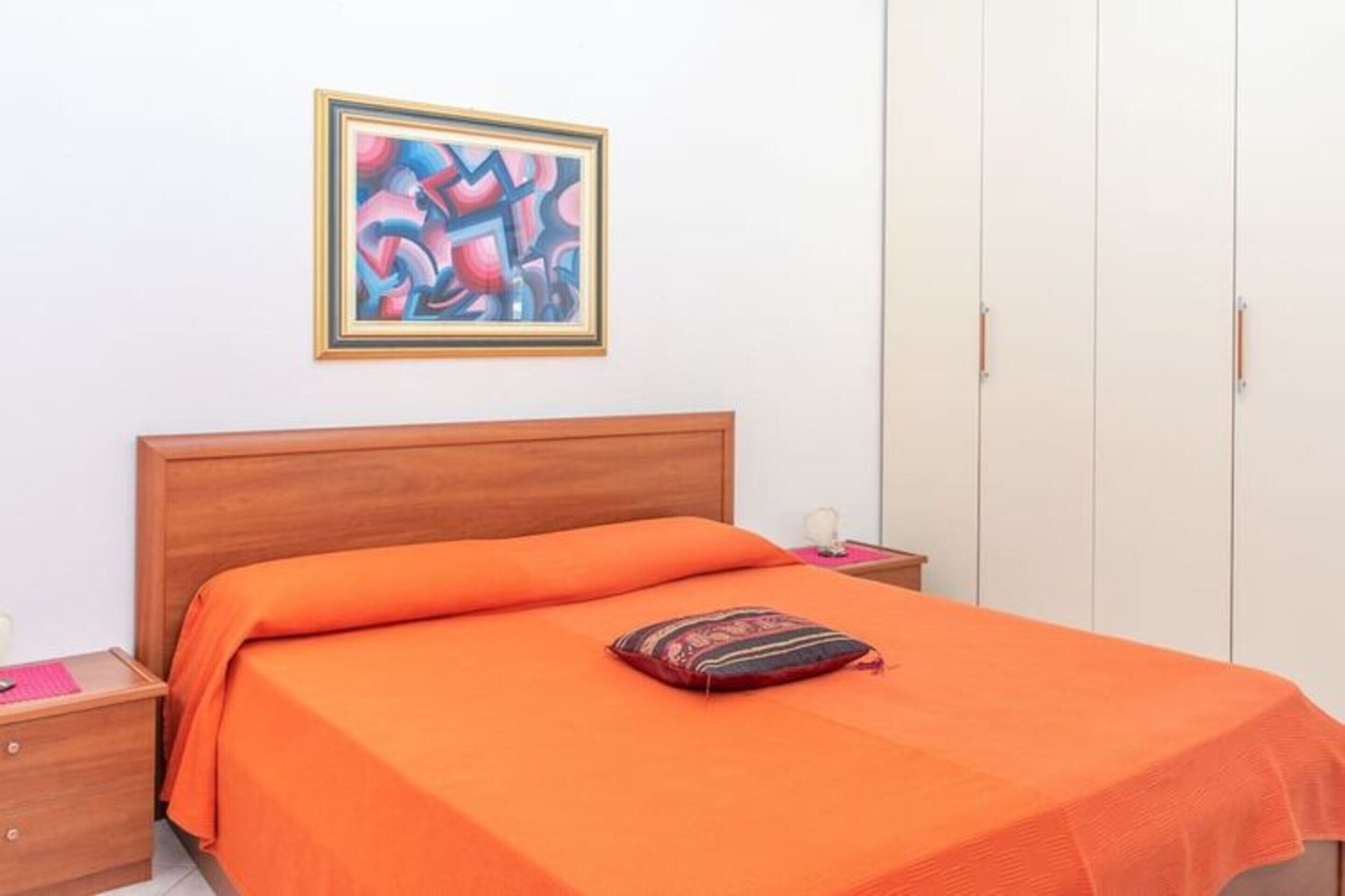 Een slaapkamer met een houten hoofdeinde, een oranje sprei, een lijst met een abstract schilderij aan de muur en een garderobe met twee deuren.