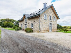 A stone house with a gravel driveway, a wooden bench, and a hedge.