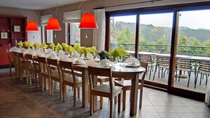 A dining area with multiple tables set for a meal, large windows with a view of a forested landscape, and red lamps above the tables.