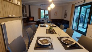 A dining area with a long table set for two, a fireplace, and a view of the outdoors through large windows.