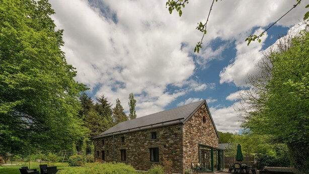 A stone building with a sloped roof, surrounded by lush greenery and a gravel driveway.