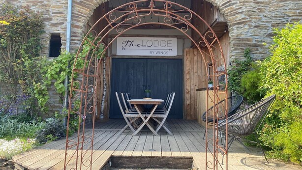 Un edificio rustico in pietra con una veranda in legno, un arco in metallo e un cartello con la scritta 'The Lodge by Wings'.