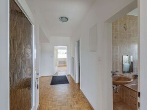 A hallway with wooden flooring, a black doormat, a white door, and a bathroom with a sink and mirror. - Nature Escape in Fuldabruck (Fuldabruck)