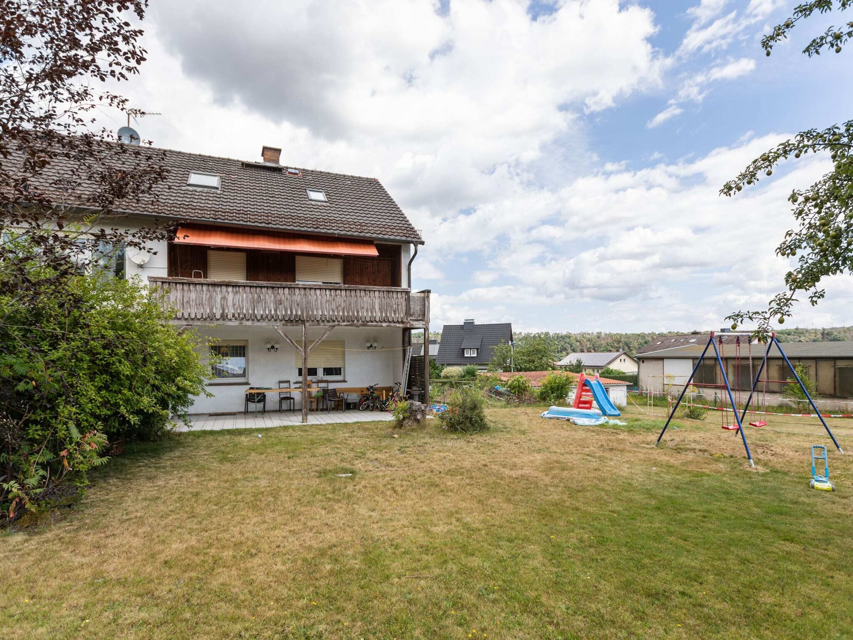 Une maison à deux étages avec un balcon, un patio aménagé avec des meubles d’extérieur et une balançoire dans le jardin.