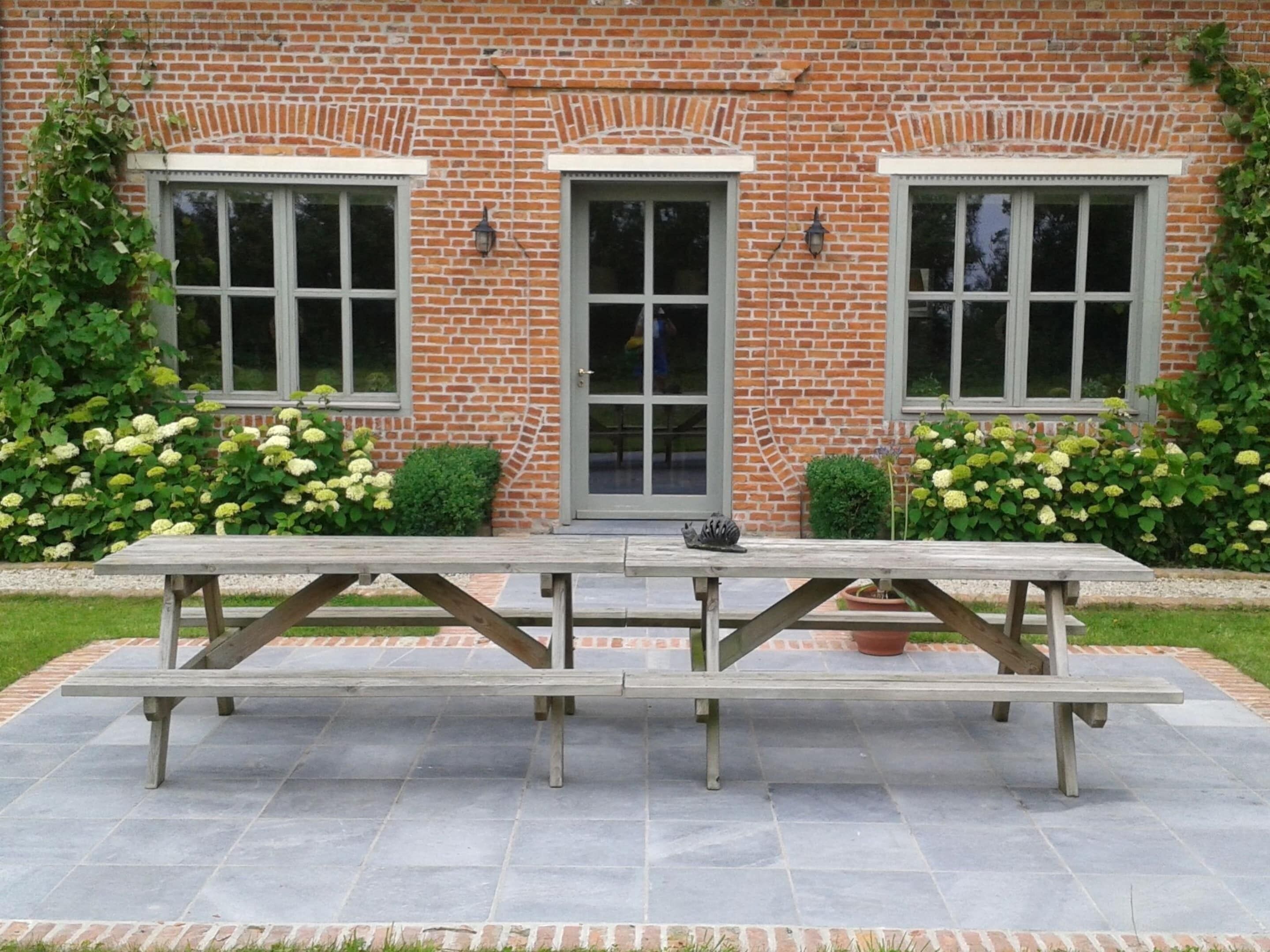 Une table de pique-nique se trouve devant un bâtiment en briques, doté de trois fenêtres et d’une porte, et est entourée de verdure et de fleurs.