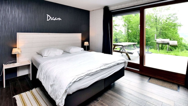 A modern bedroom with a large bed, a nightstand, a wall-mounted lamp, and a view of the outdoors through a sliding glass door. - Luxurious Chalet in Beauraing With Sauna (Beauraing)