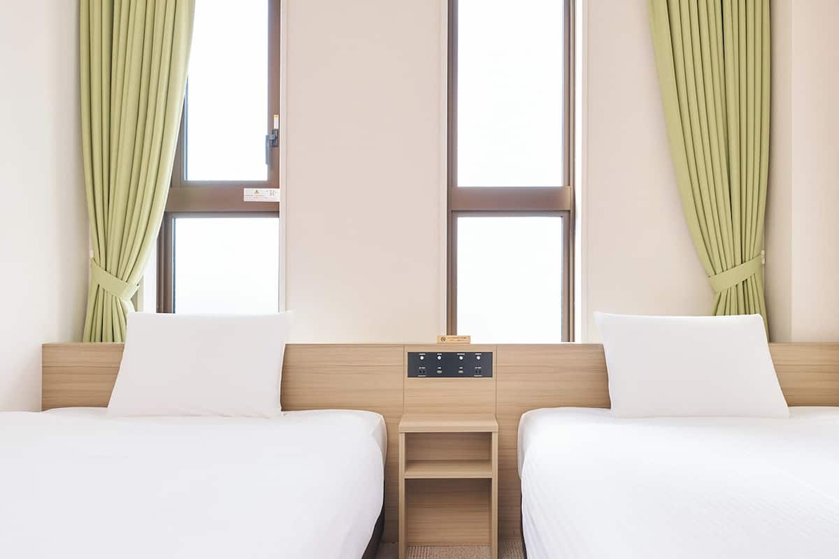 Ein Hotelzimmer mit zwei Einzelbetten, einem Holznachttisch, einer Steuerung und grünen Vorhängen.