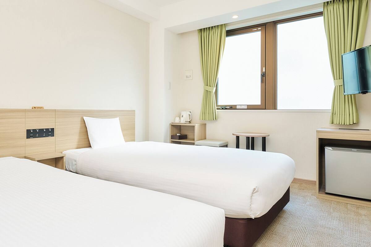 Ein Hotelzimmer mit zwei Betten, einem Fernseher, einer kleinen Kühlbox und einem Fenster mit Vorhängen.
