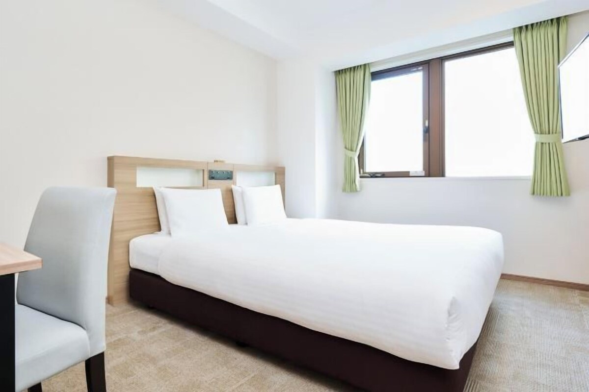 Ein Hotelzimmer mit Bett, Stuhl, Nachttisch und einem Fenster mit Vorhängen.