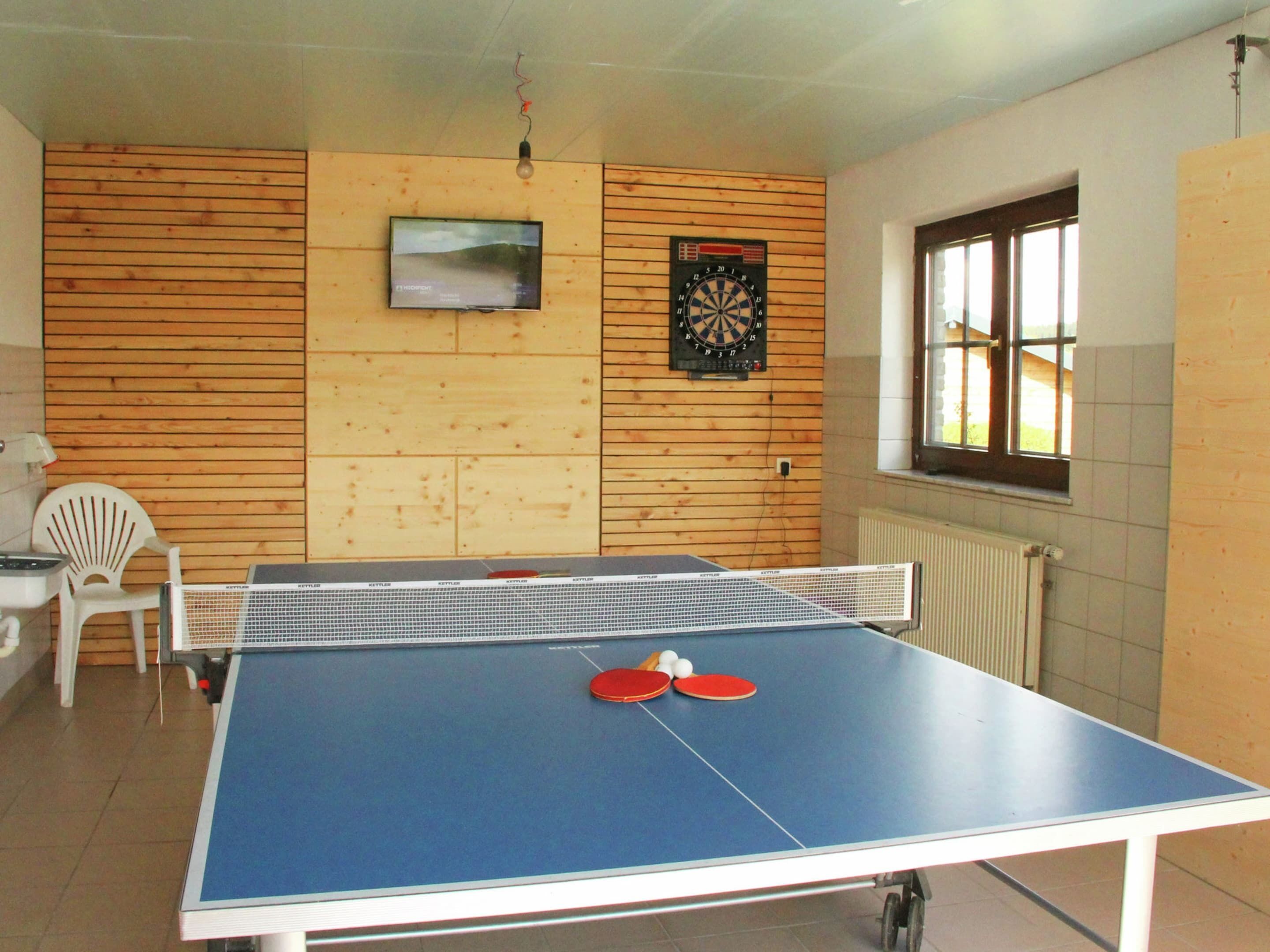 Egy pingpongasztal, két piros ütővel és két fehér labdával, egy dartstábla, egy televízió és egy ablak, amelyen zsaluzi van.