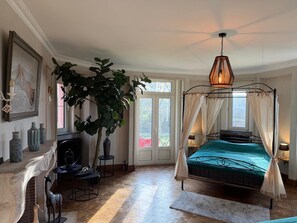Une chambre à coucher avec un lit, une grande plante, une cheminée et une vue sur l’extérieur.