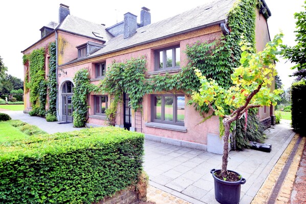 Une maison à deux étages avec une façade en briques, de grandes fenêtres et un jardin bien entretenu agrémenté d’une haie et d’un arbre en pot.