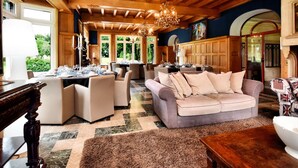 A spacious living room with a high ceiling, wooden paneling, and a chandelier. There is a dining area with tables set for a meal. - Magnificent Luxury Chateau in Vast Garden with Sauna & Hot Tubs (Chiny)