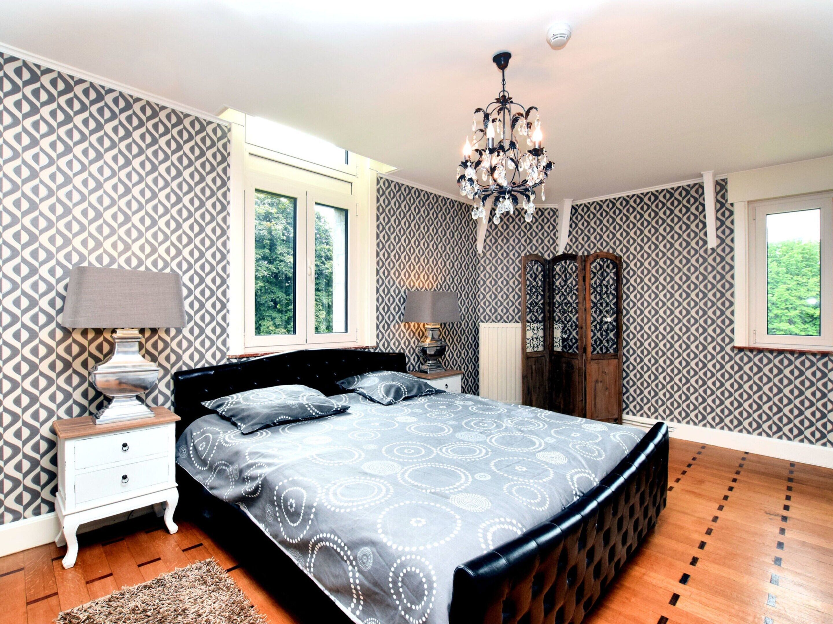 A bedroom with a large bed, two bedside tables, a dresser, a chandelier, and patterned wallpaper.