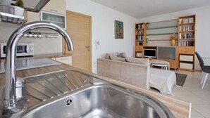 Eine moderne Küche mit Spüle aus Edelstahl, einer Mikrowelle und einer Holzarbeitsplatte. Im Wohnbereich befindet sich ein beiges Sofa und ein Bücherregal.