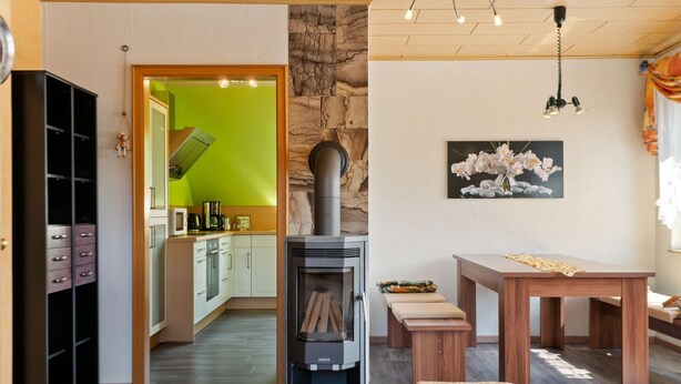 A modern kitchen with a green wall, a stone wall, and a Justus wood stove. There is a dining area with a table and bench.