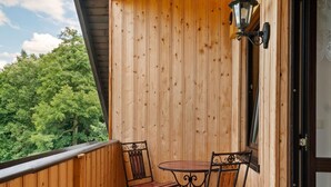 A wooden balcony with two metal chairs and a small table, overlooking a forested area.