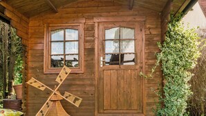 A wooden cabin with a pitched roof, two windows with lace curtains, and a windmill model in front.