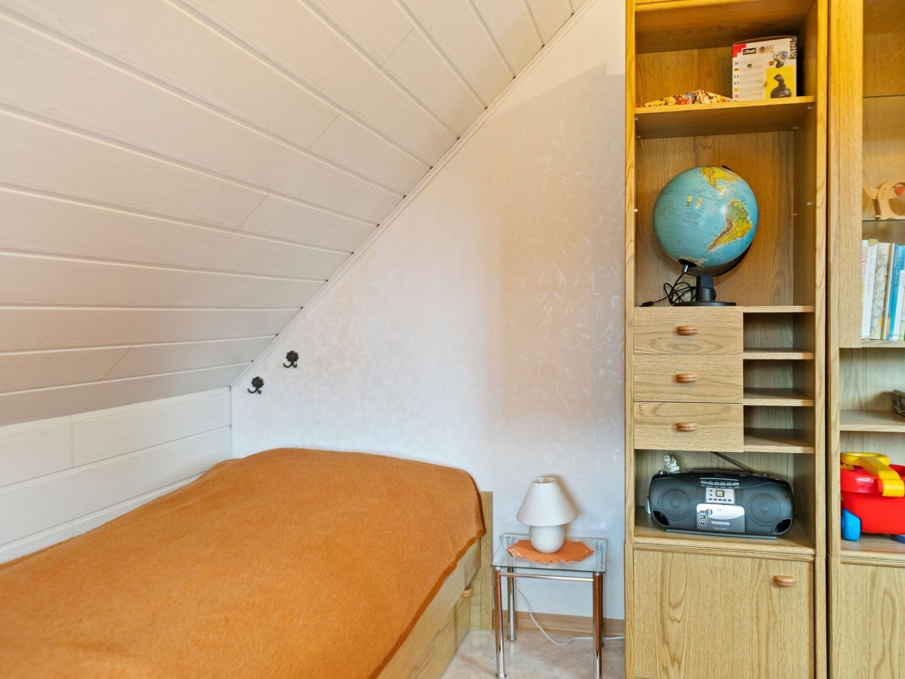 A small bedroom with a single bed, a nightstand with a lamp, a bookshelf with books and a globe, and a wooden shelf with toys.