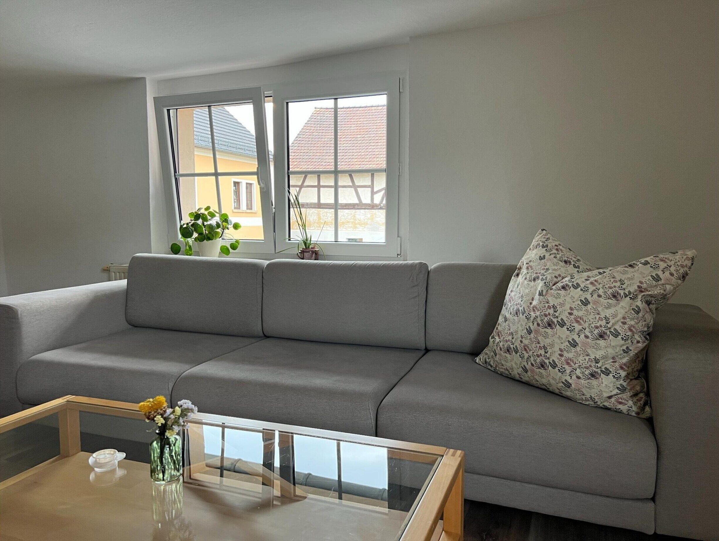 A living room with a grey sofa, a glass coffee table, and a window with a view of buildings.