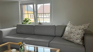 A living room with a grey sofa, a glass coffee table, and a window with a view of buildings.