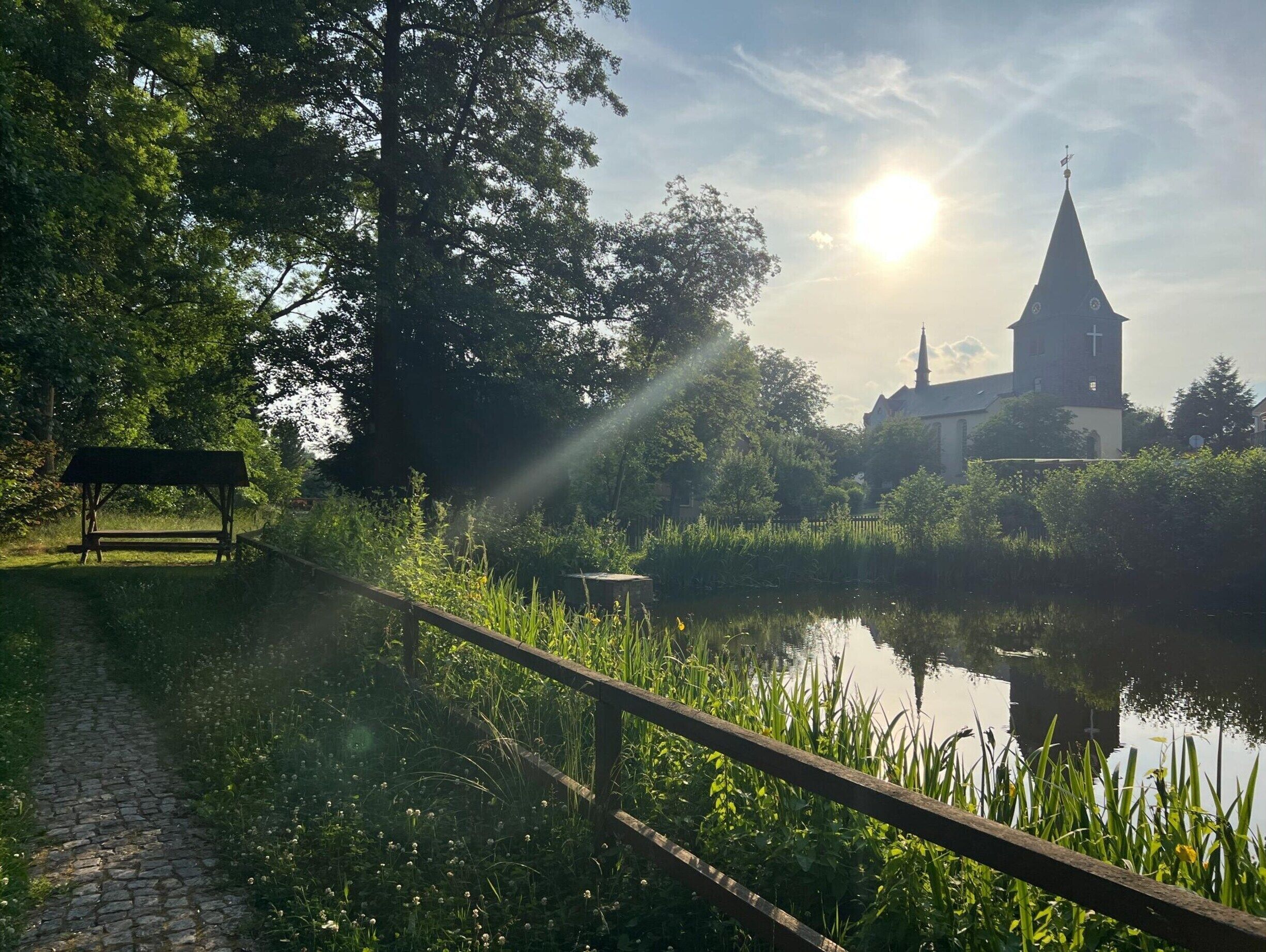 Un sendero tranquilo a la orilla del río con un banco, abundante vegetación y una iglesia al fondo.