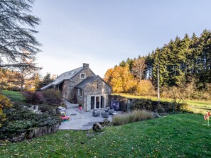A stone house with a sloped roof, a patio with outdoor furniture, and a garden with trees and shrubs. - Luxurious Farmhouse Near Forest in Anhee (Anhée)