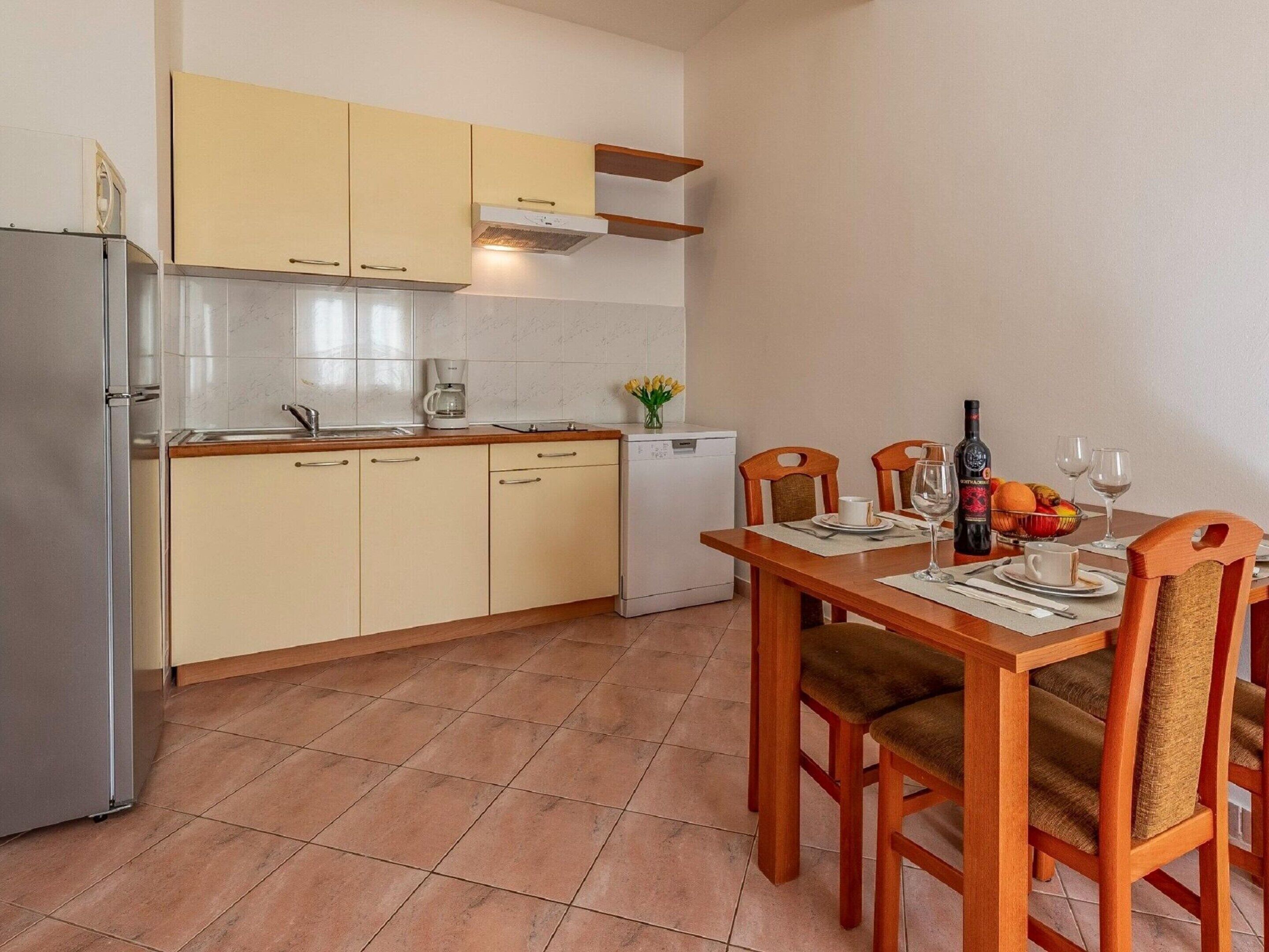 Cocina compacta con refrigerador, cafetera y un lavamanos pequeño. Hay una mesa de comedor con sillas y una botella de vino.