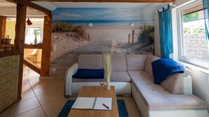 A living room with a beach mural, a sofa, a coffee table, and a dining area with a brick wall and wooden beams. - Pleasant Apartment in Veckenstedt near Ilsenburg (Nordharz)