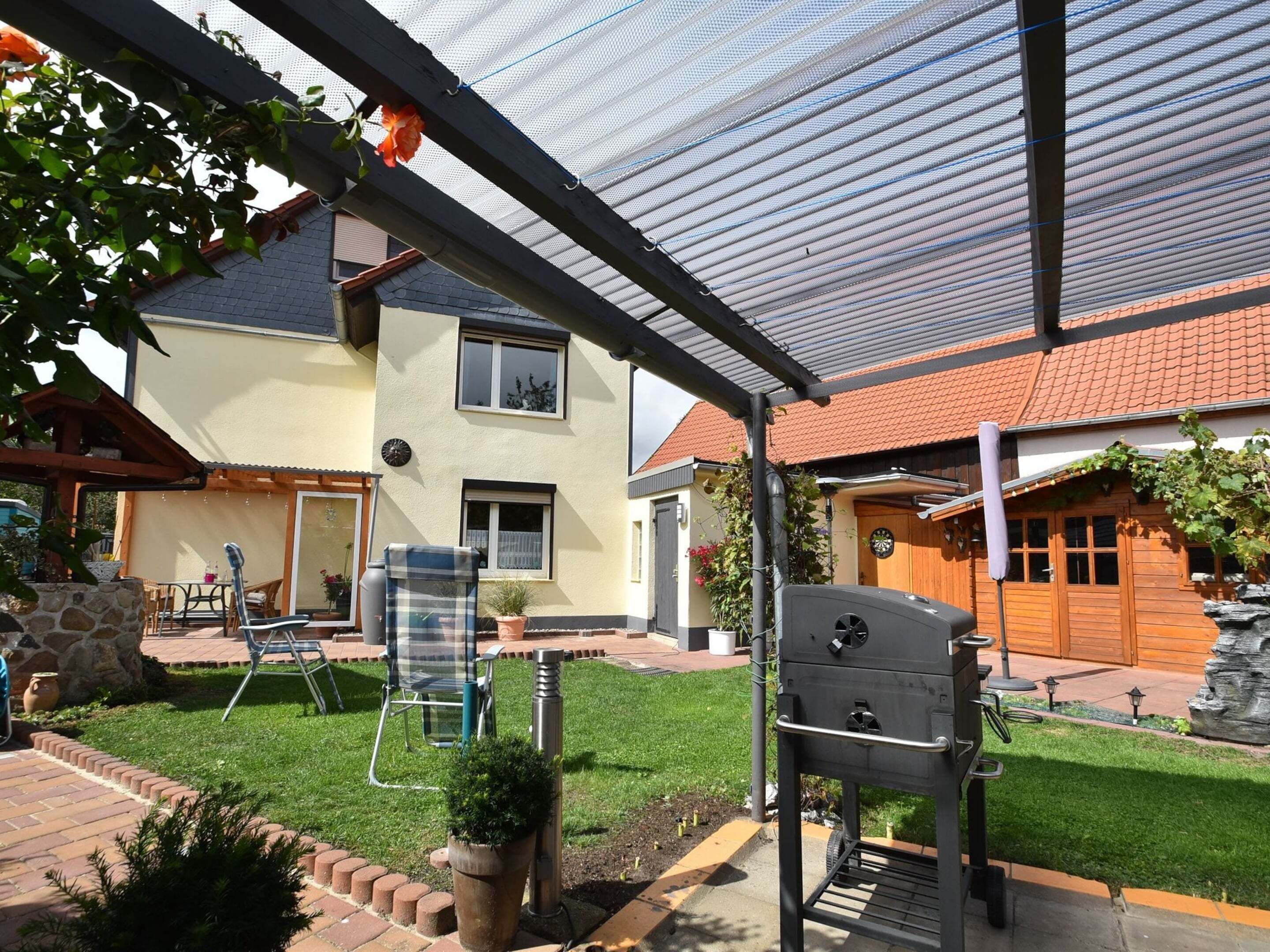 A covered patio area with a grill, outdoor furniture, and a house in the background.