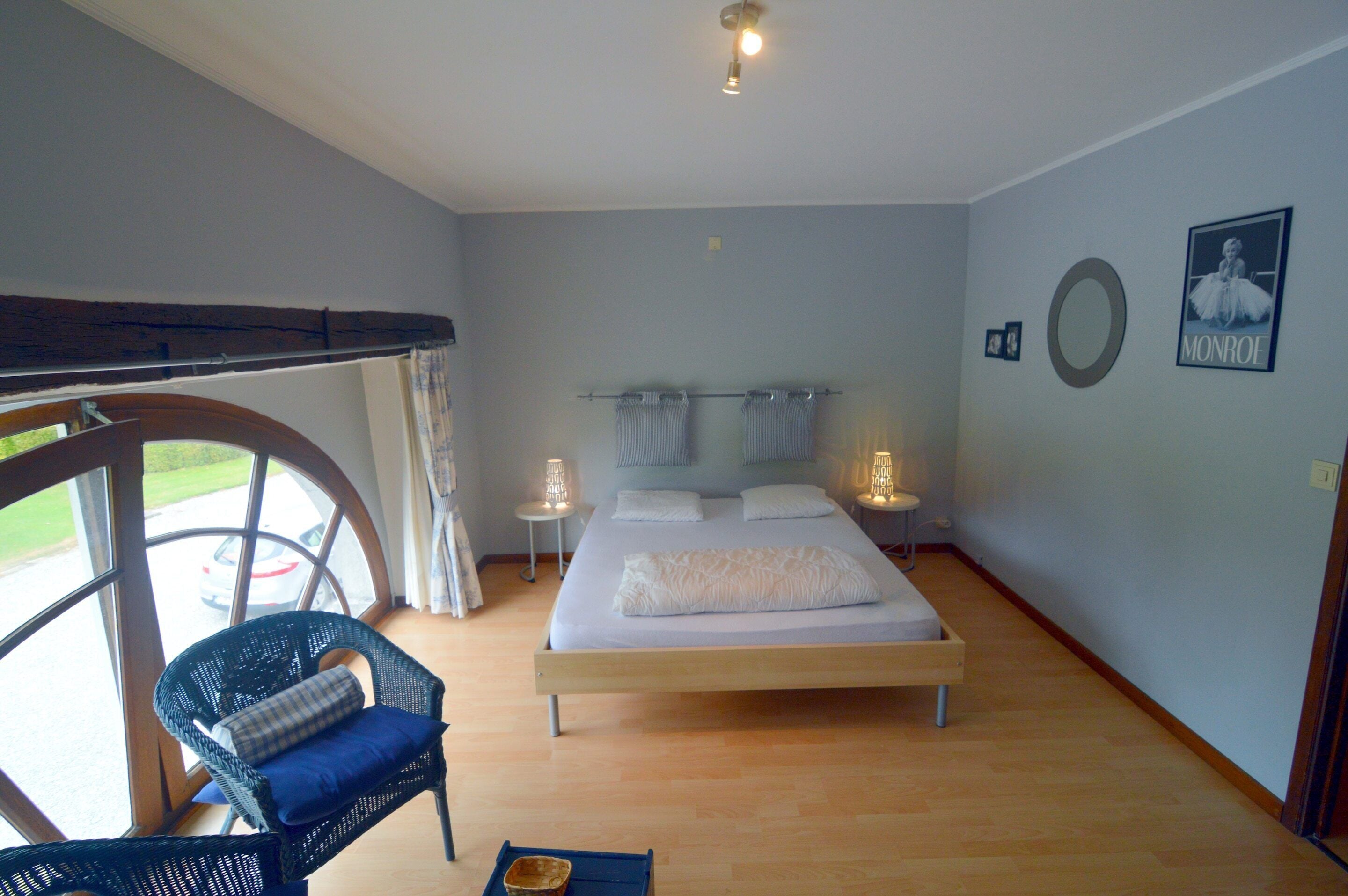 A bedroom with a bed, a wicker chair, a circular window, and a wall-mounted picture.