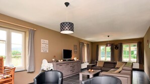 A spacious living area with a sofa set, two armchairs, a coffee table, and a television. There are windows with curtains, a wooden chair, and a small table with a vase. - Holiday Home in Hastiere Near Forest (Hastiere)