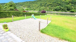 A playground with a slide and a fenced area, surrounded by lush greenery and a lake. - Holiday Home in Hastiere Near Forest (Hastiere)