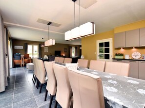A dining area with a long table, beige chairs, and a chandelier. There is a kitchen area with cabinets and a clock on the wall. - Holiday Home in Hastiere Near Forest (Hastiere)
