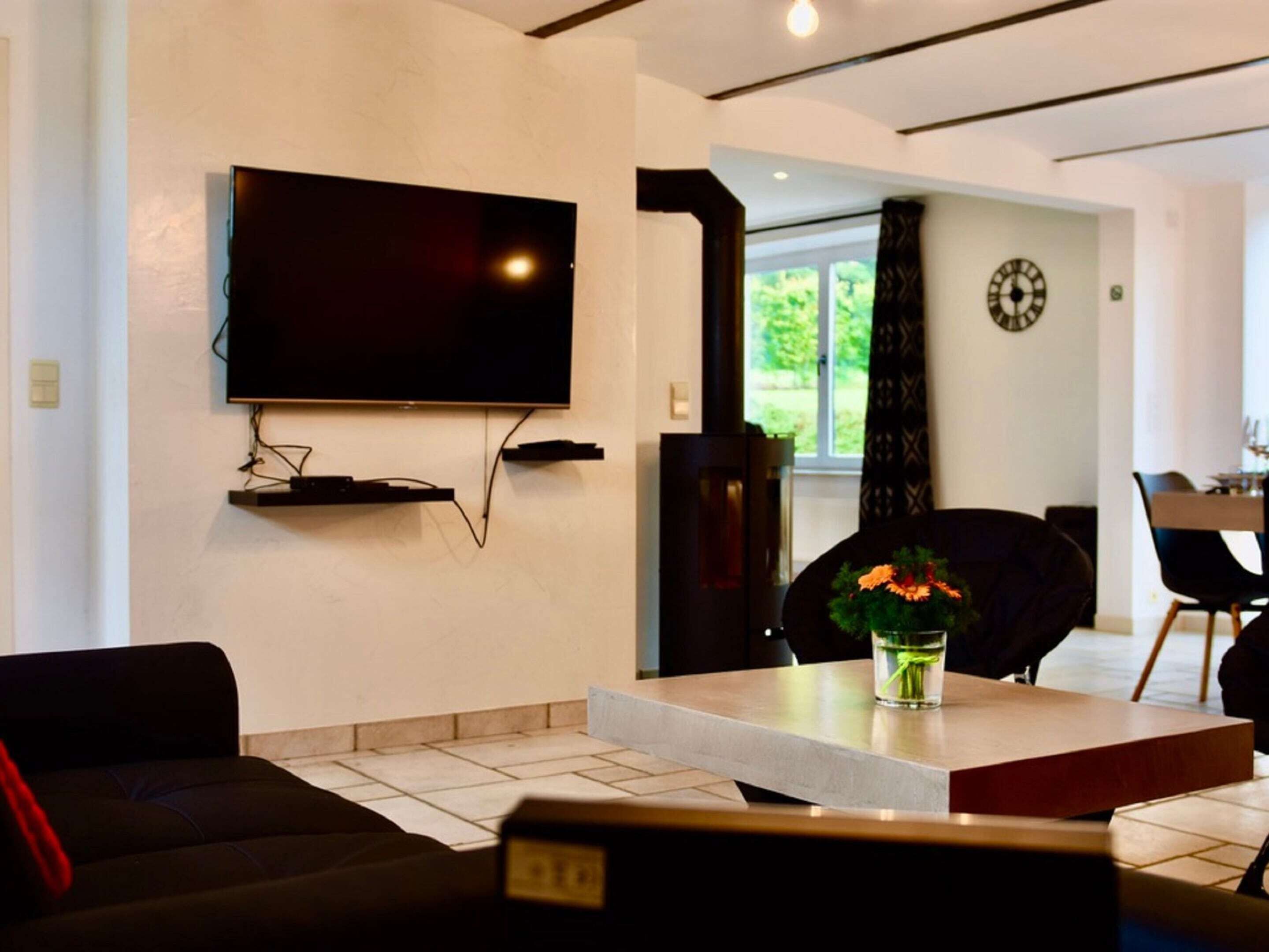 A modern living room with a flat-screen TV mounted on the wall, a dining area with a table and chairs, and a large window with a view of greenery.