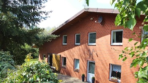 A red brick building with multiple windows, a door, and a small garden area with potted plants. - Cozy Apartment With Pool & Forest (Kirchdorf)