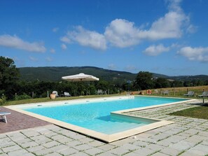 A rectangular swimming pool with a clear blue water surface, surrounded by a paved deck with grass inlays. There are lounge chairs and a white umbrella providing shade. The background features a hilly landscape under a clear sky.