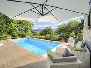 Ein Swimmingpool mit Holzterrasse, Gartenmöbeln und Blick auf Grünflächen und Berge.