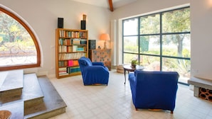 A spacious living room with a large window, a bookshelf, two blue armchairs, and a small table.