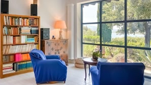 A room with a large window, a bookshelf, two blue armchairs, a small table with a lamp, and a radiator.