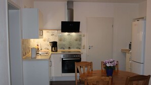 A compact kitchen with white cabinets, a built-in oven, a microwave, and a black TV above the oven. There is a dining table with chairs and a blue vase with flowers. - Chicke Ferienwohnung in Struppen (Struppen)