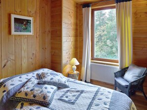 Quarto de cabana de madeira com cama, uma cadeira, janela com cortinas e um quadro na parede.