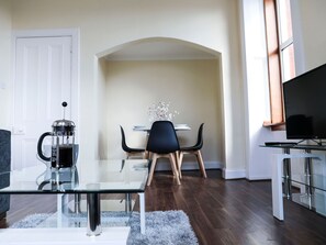 A modern living room with a glass coffee table, a black sofa, a coffee maker, a dining table with chairs, and a TV on a stand.