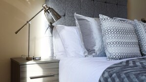 A neatly made bed with a grey headboard, a bedside lamp, and a wooden nightstand with drawers.