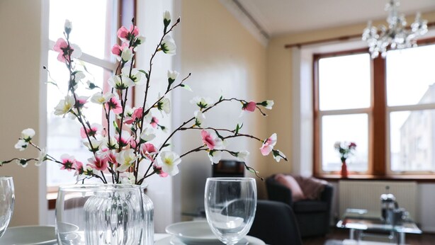 A dining table set with plates, glasses, and cutlery, a vase with flowers, and a sofa in the background.